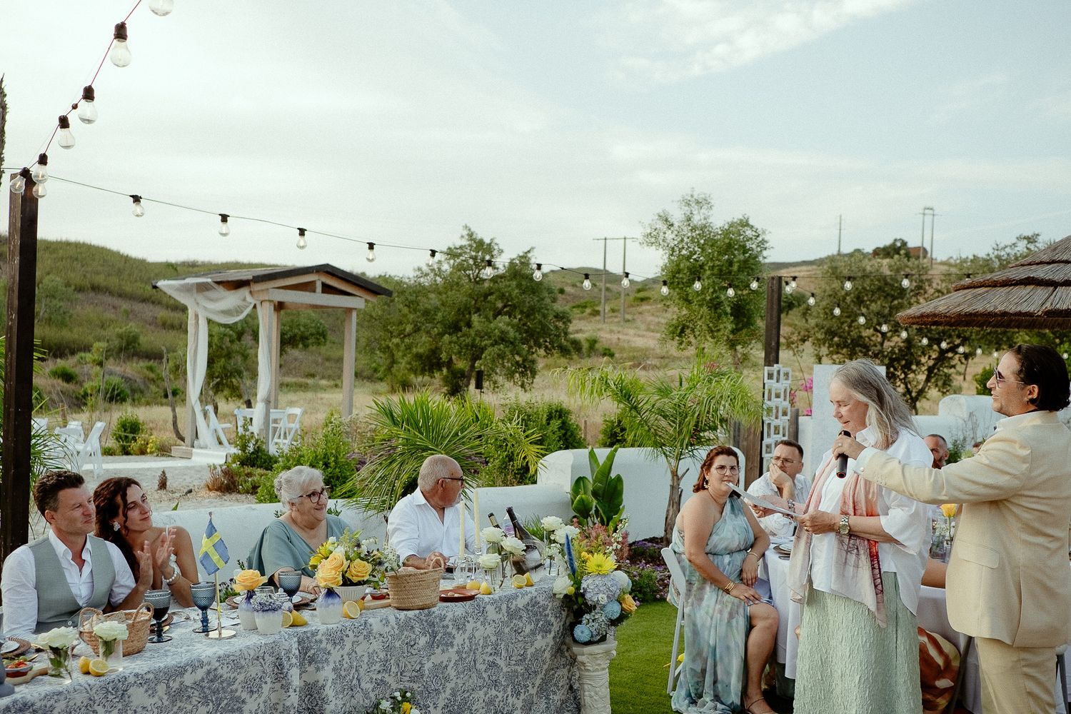 Outdoor wedding reception with guests seated at decorated tables, string lights, and rolling hills backdrop.