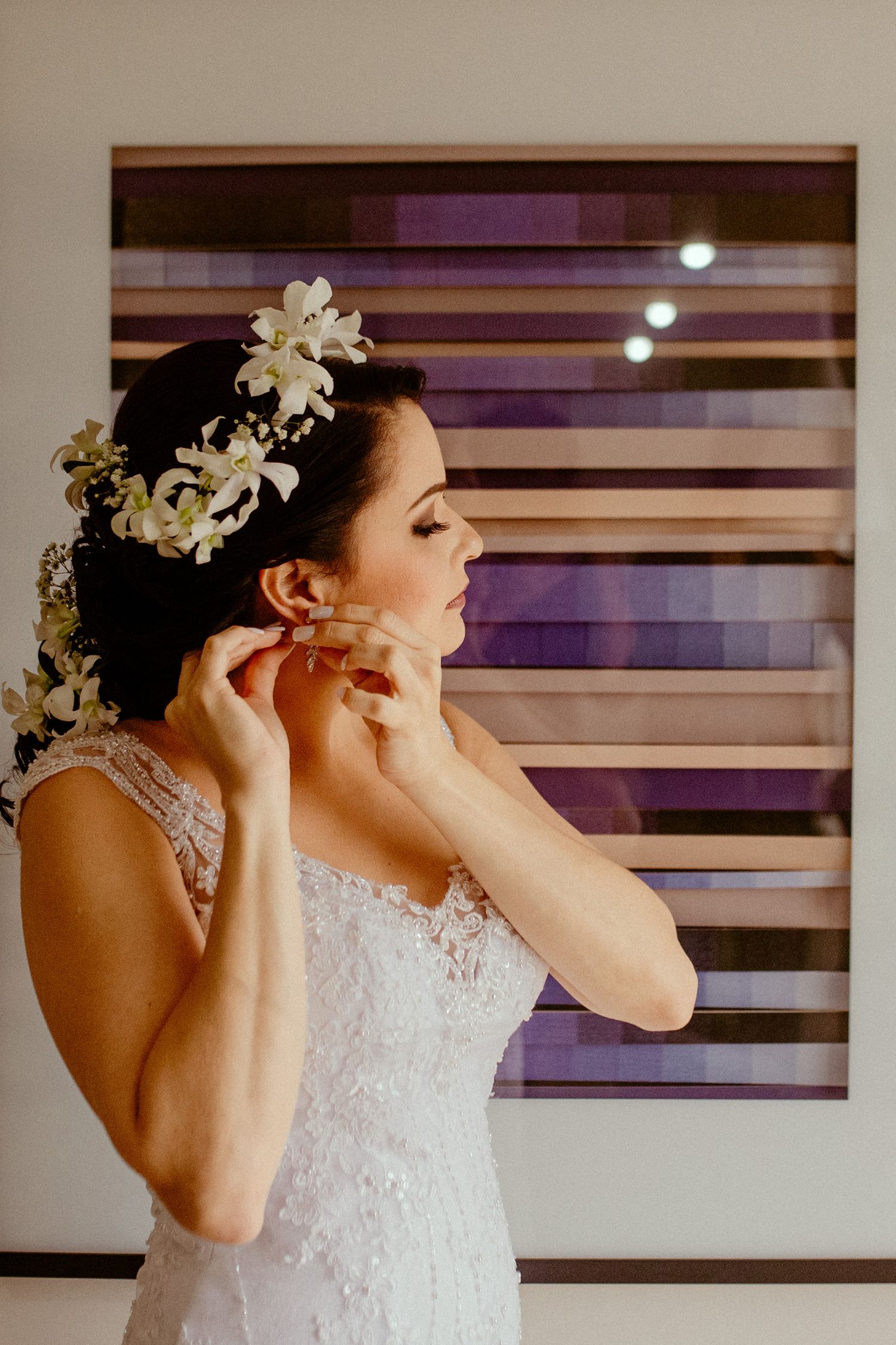A bride in a white lace gown adjusts her earring while wearing a delicate floral crown against a striped wall background.