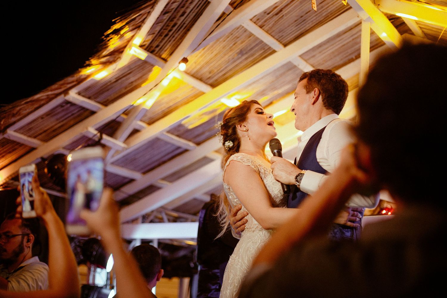 A couple dancing together at an evening wedding reception under warm lighting while guests capture the moment on phones.