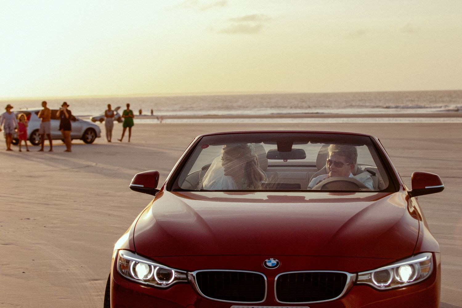 Red BMW convertible parked on beach at sunset with people walking in background.