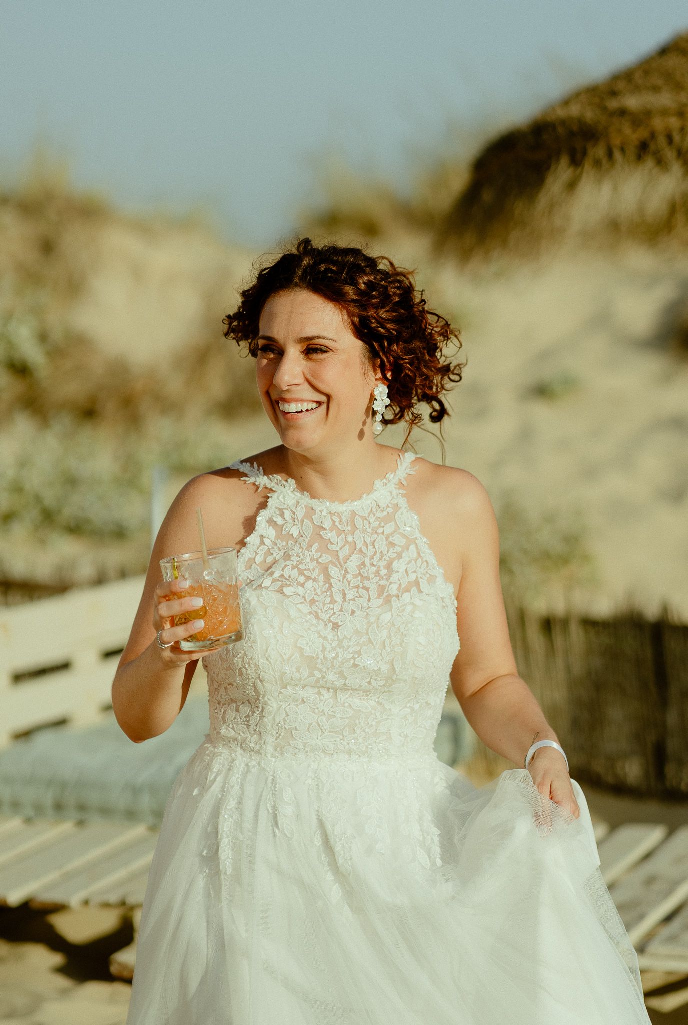 Smiling bride in white lace halter gown holding a drink outdoors on a sunny beach boardwalk.