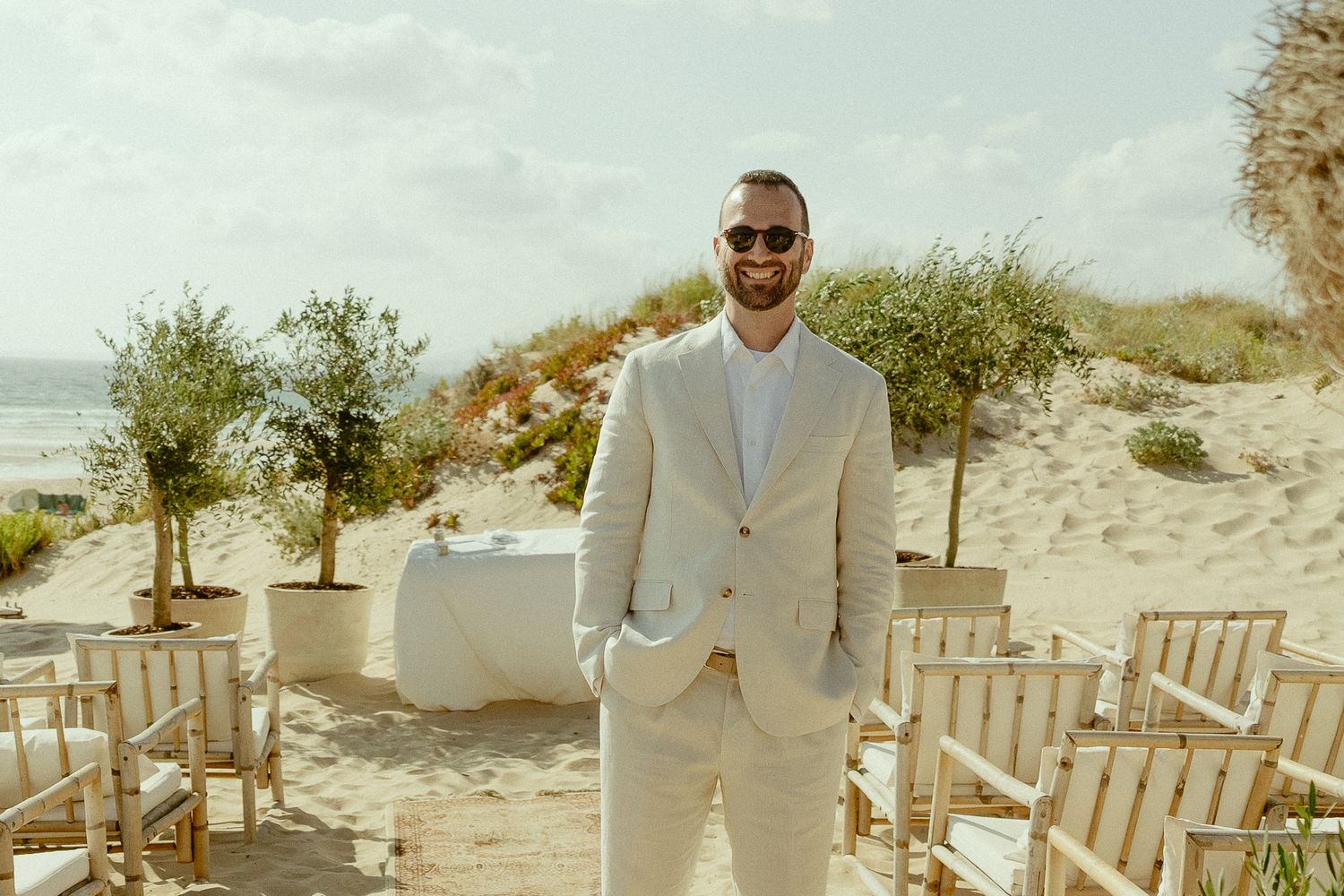 Man in beige suit and sunglasses smiling at beach wedding ceremony with white chairs and olive trees.