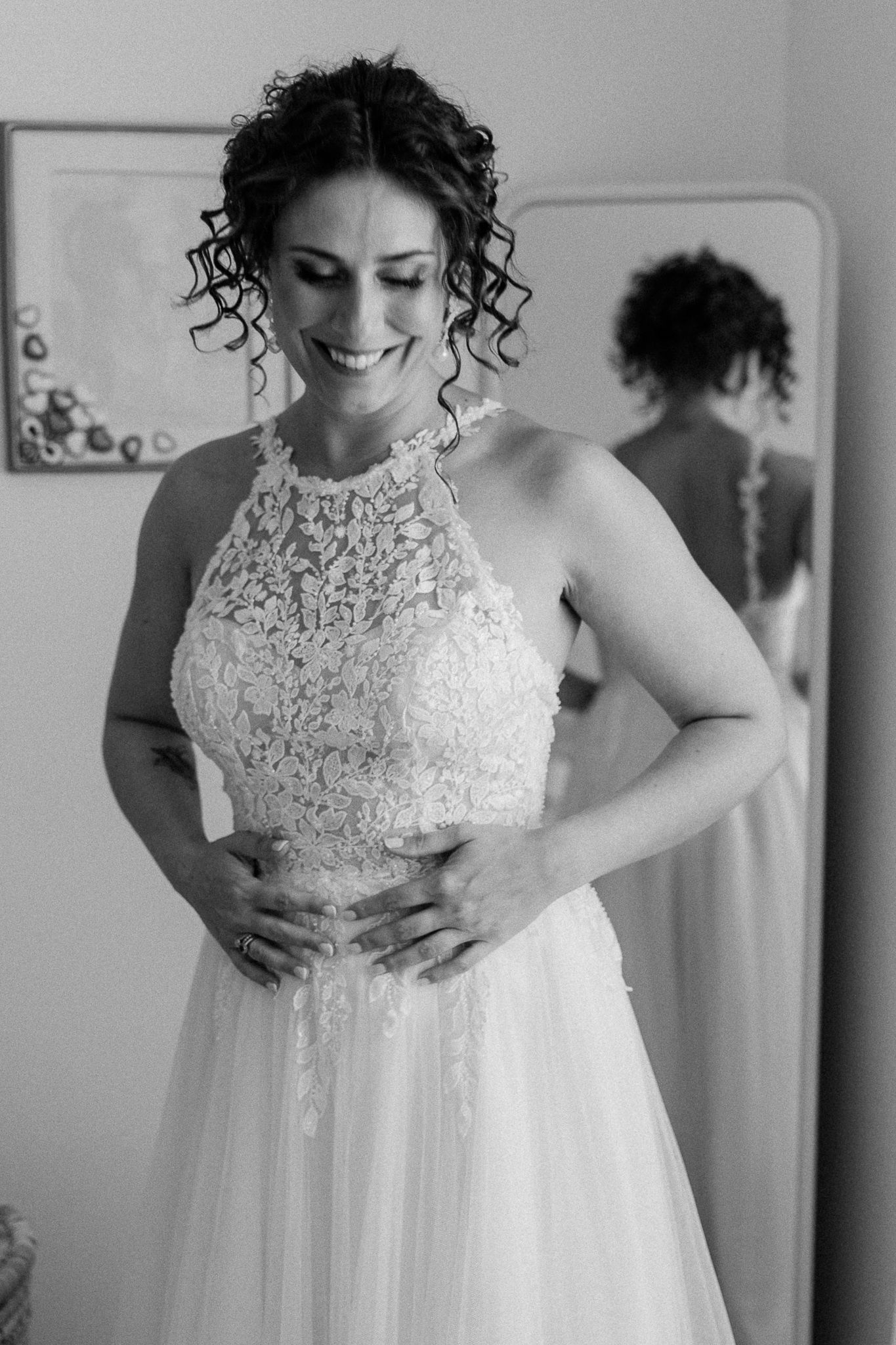 Black and white photo of a smiling bride in a lace halter wedding gown, with her reflection visible in a mirror behind her.