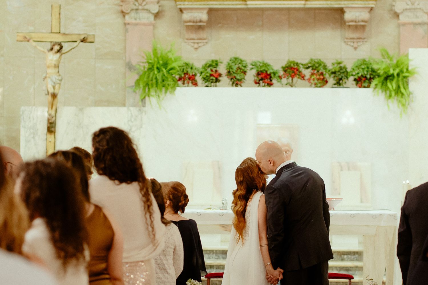 Bride and groom kneeling at church altar during wedding ceremony with floral decorations and crucifix.