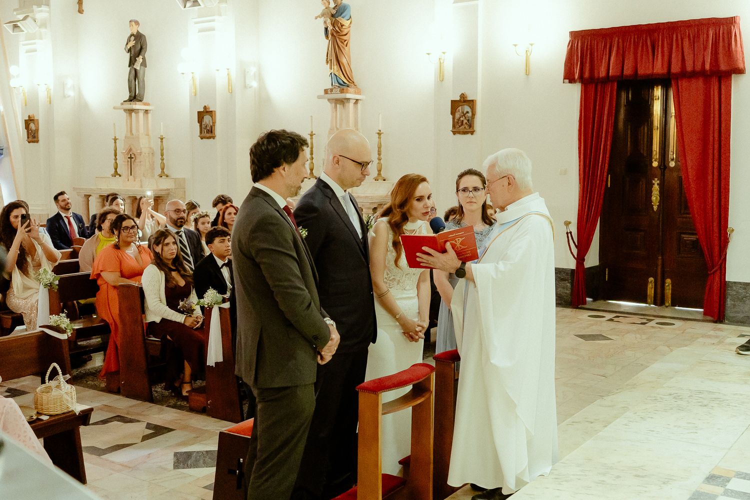 Couple exchanging vows at a Catholic church wedding ceremony with priest and guests seated in pews.