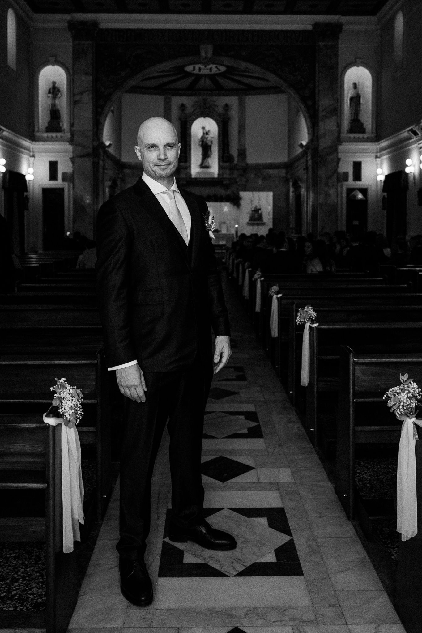 Groom in black suit standing in church aisle with decorated pews, black and white wedding photo.