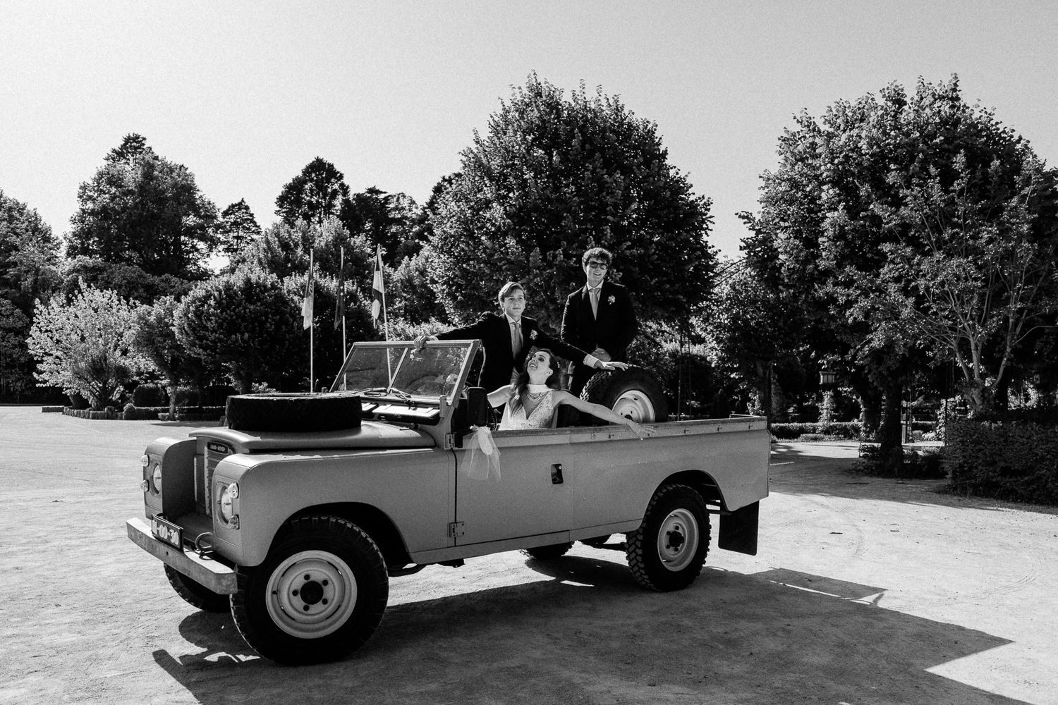 Black and white photo of a wedding party posing in a vintage Land Rover convertible surrounded by lush trees.