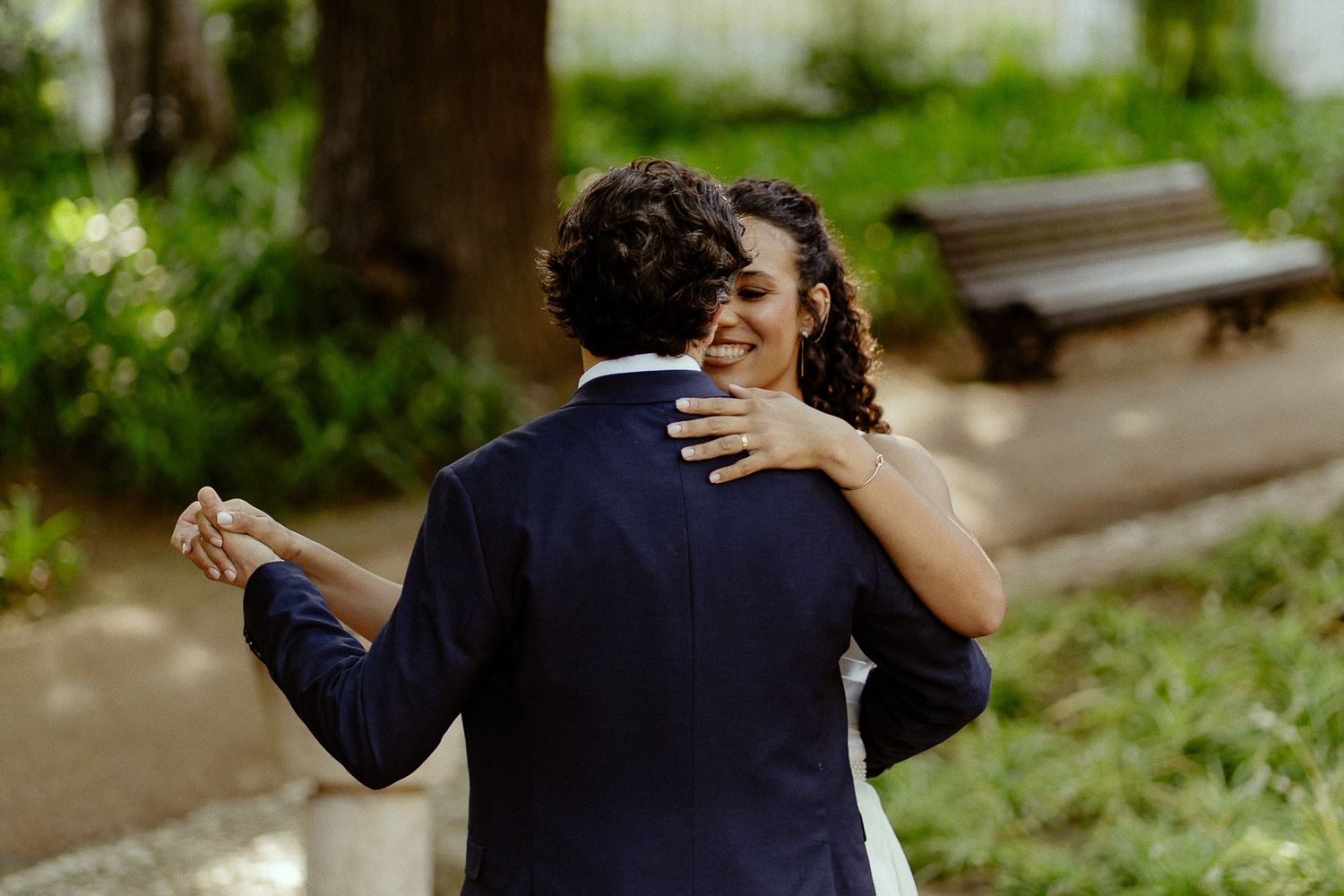 A joyful bride and groom share a dance outdoors in a lush green park on their wedding day.