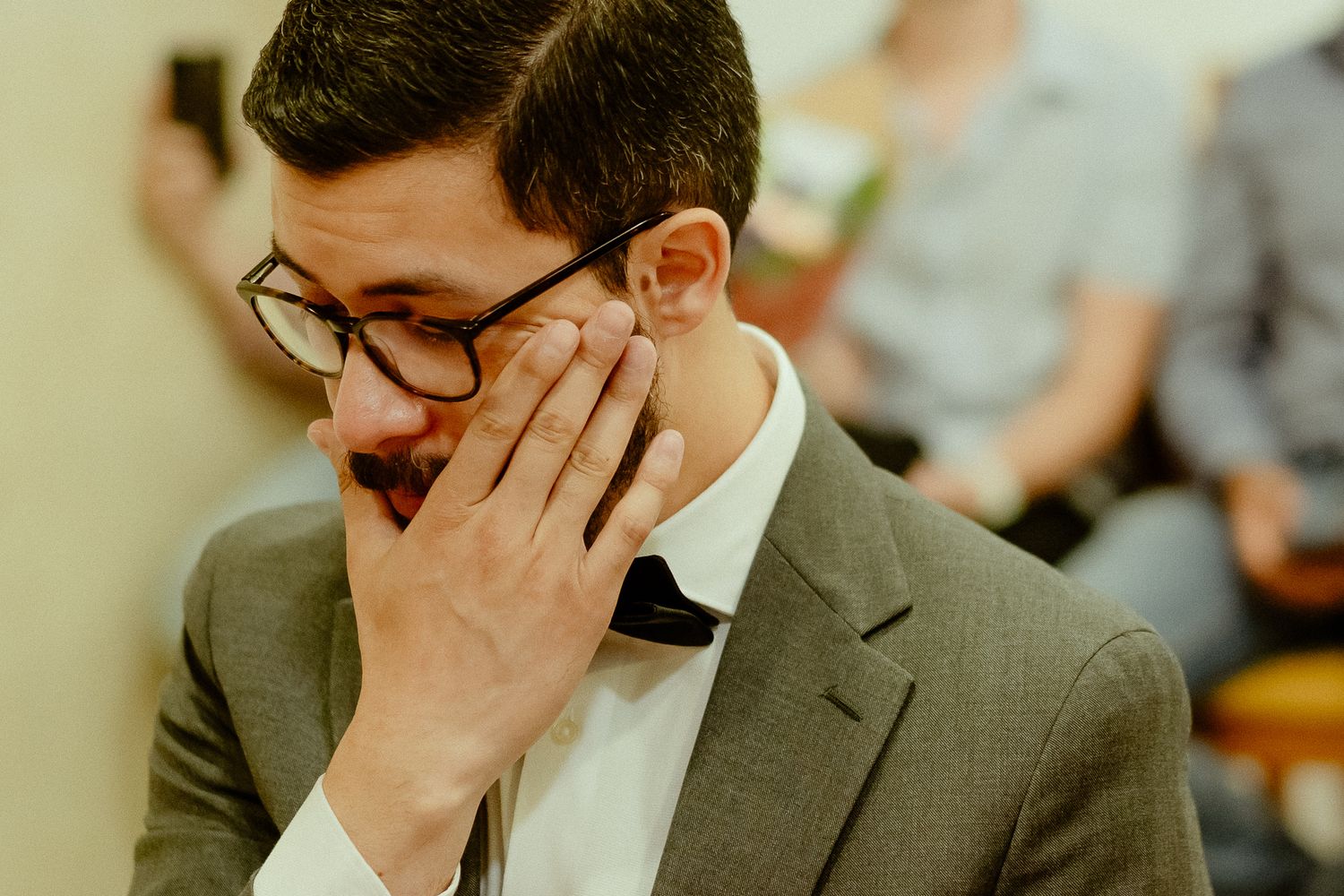 Emotional man in glasses and suit covering mouth with hand during a heartfelt moment at a formal event.