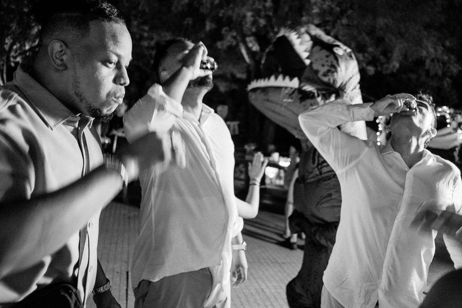 Black and white photo of men in dress shirts raising drinks at an outdoor evening celebration or wedding reception.