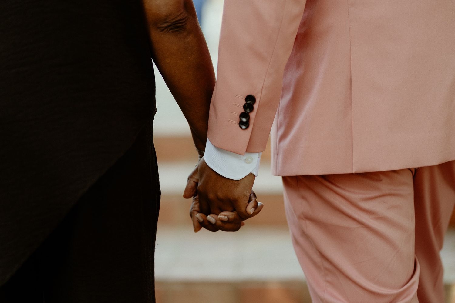 Two people holding hands, one wearing a pink suit and the other in a black outfit, symbolizing love and togetherness.