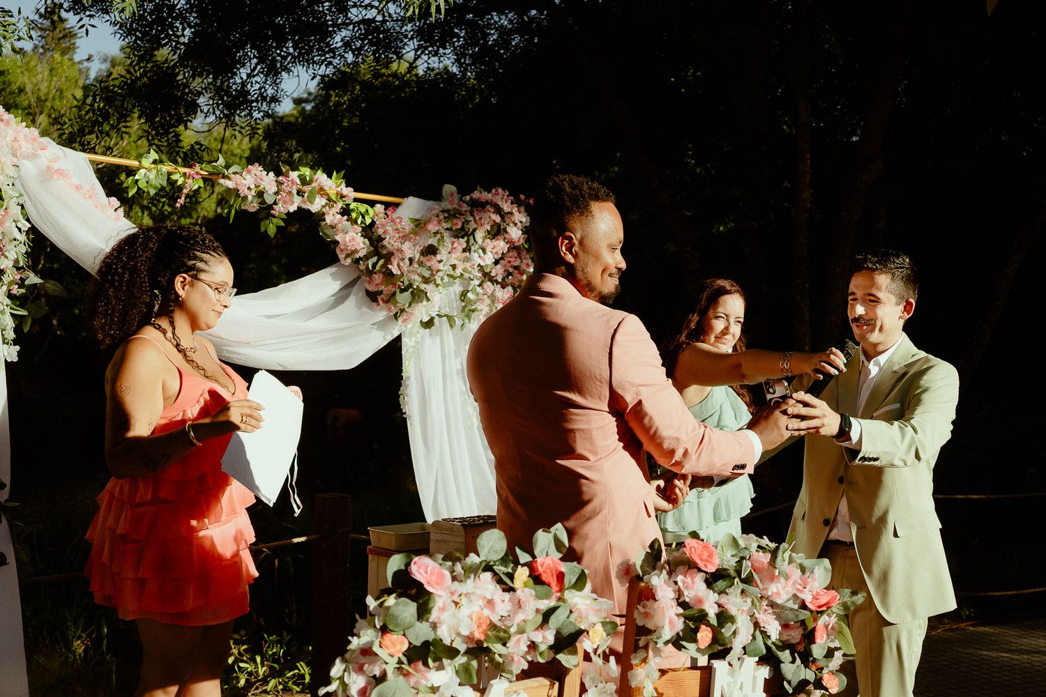 Two grooms exchange rings at an outdoor wedding ceremony beneath a floral arch with pink blooms.