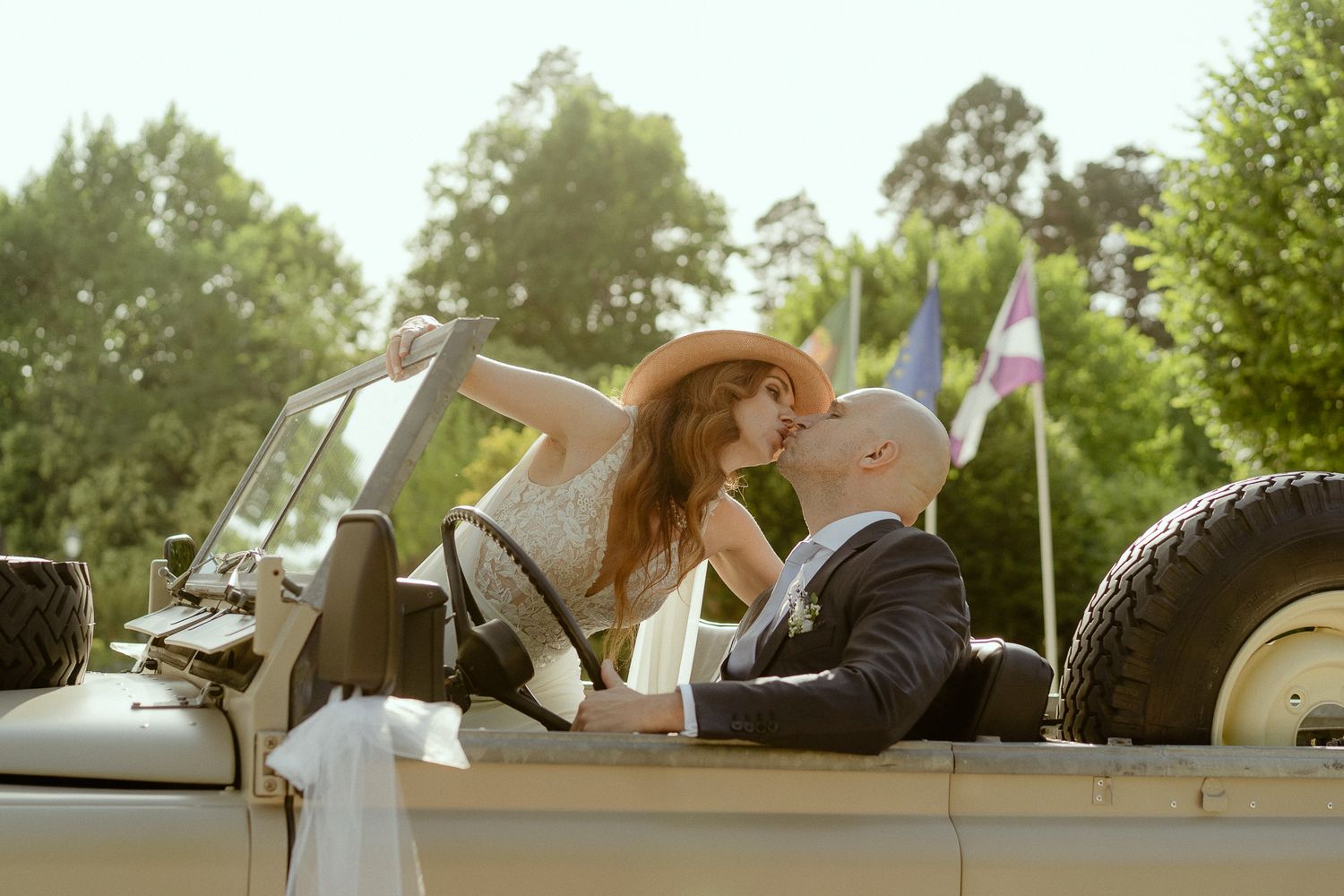 A couple shares a romantic kiss in a vintage convertible car surrounded by lush green trees and colorful flags.