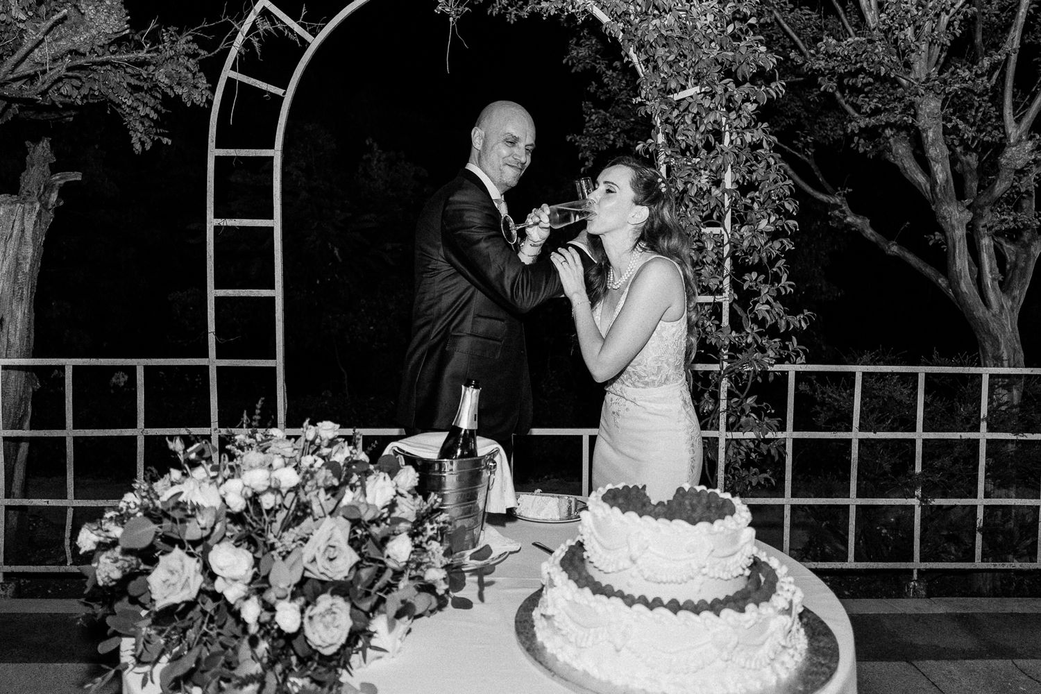 Bride and groom cutting wedding cake outdoors at night, black and white photo with floral arrangements.