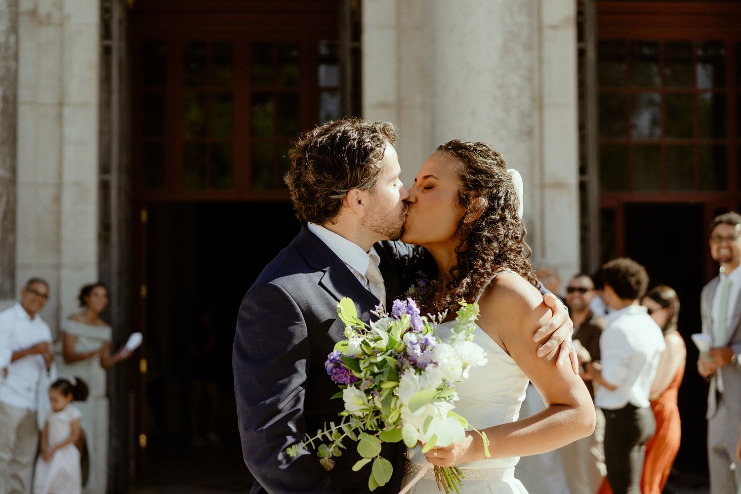 A newlywed couple shares a romantic kiss outside a grand building, bride holding a purple and white bouquet.