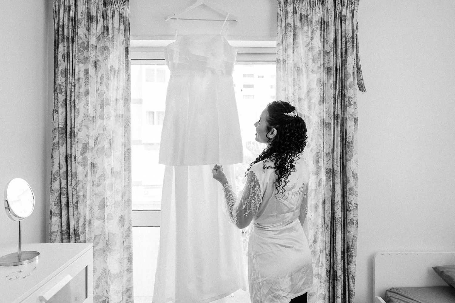 Bride in robe admiring her white wedding dress hanging by a window in a black and white photo.