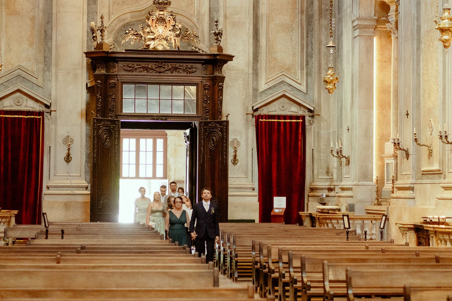 Wedding procession walking down the aisle of an ornate baroque church with wooden pews and red curtains.