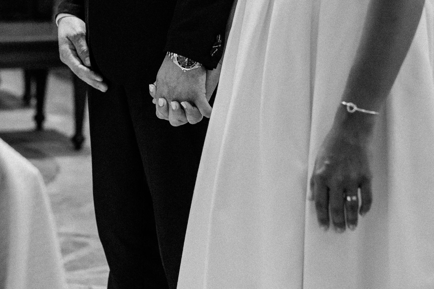 Black and white wedding photo of bride and groom holding hands, showcasing elegant attire and jewelry.