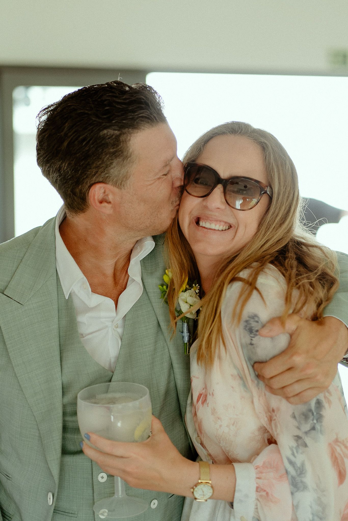 A man in a sage suit kisses a smiling woman in a floral dress wearing sunglasses at a wedding celebration.