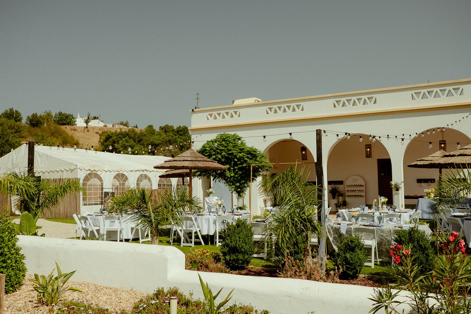 Elegant outdoor Mediterranean restaurant with white chairs, tables, and arched building surrounded by lush greenery under clear sky.