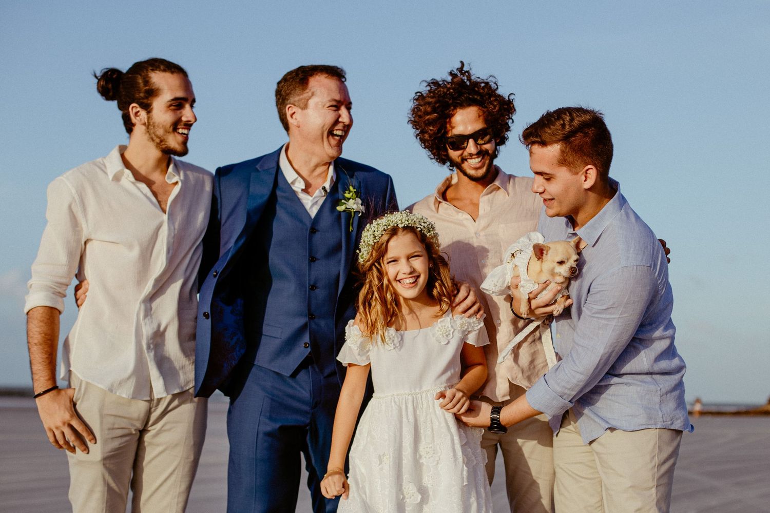 Group portrait of wedding party members smiling together on beach at sunset.