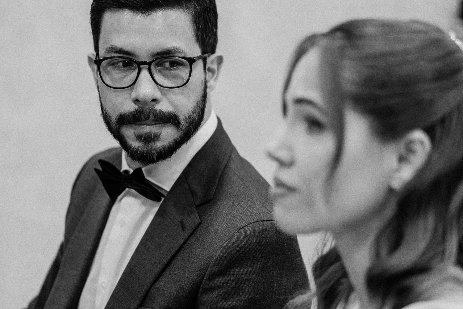 Black and white wedding photo of groom in bow tie and glasses gazing at bride in soft focus foreground.