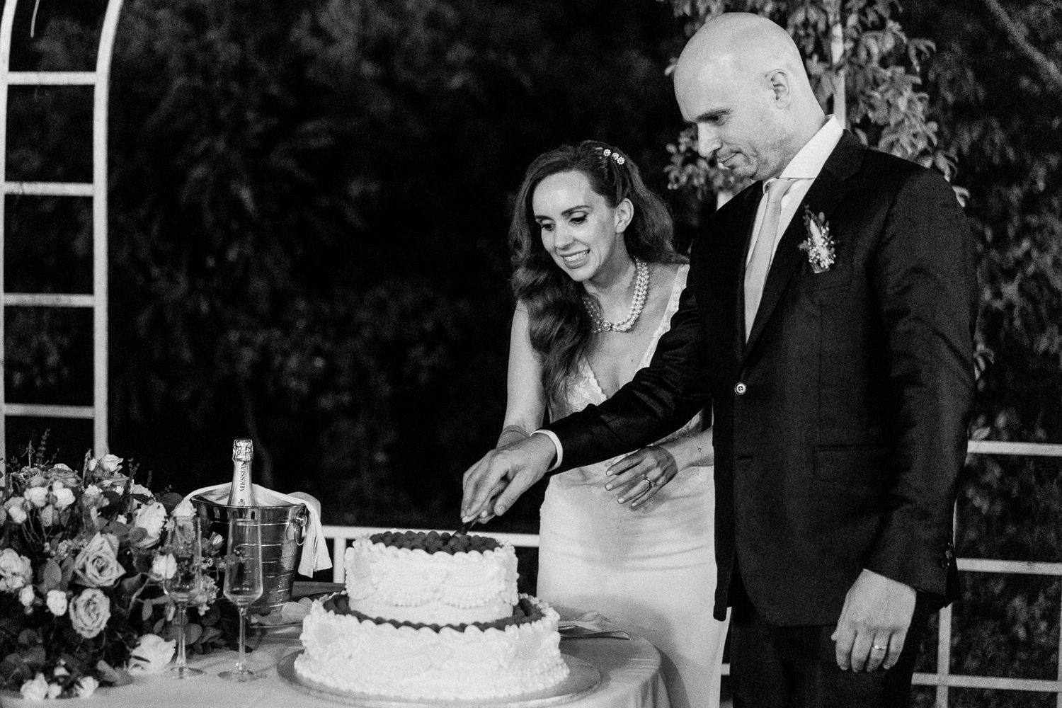 Black and white photo of a bride and groom cutting their wedding cake at an elegant outdoor reception.