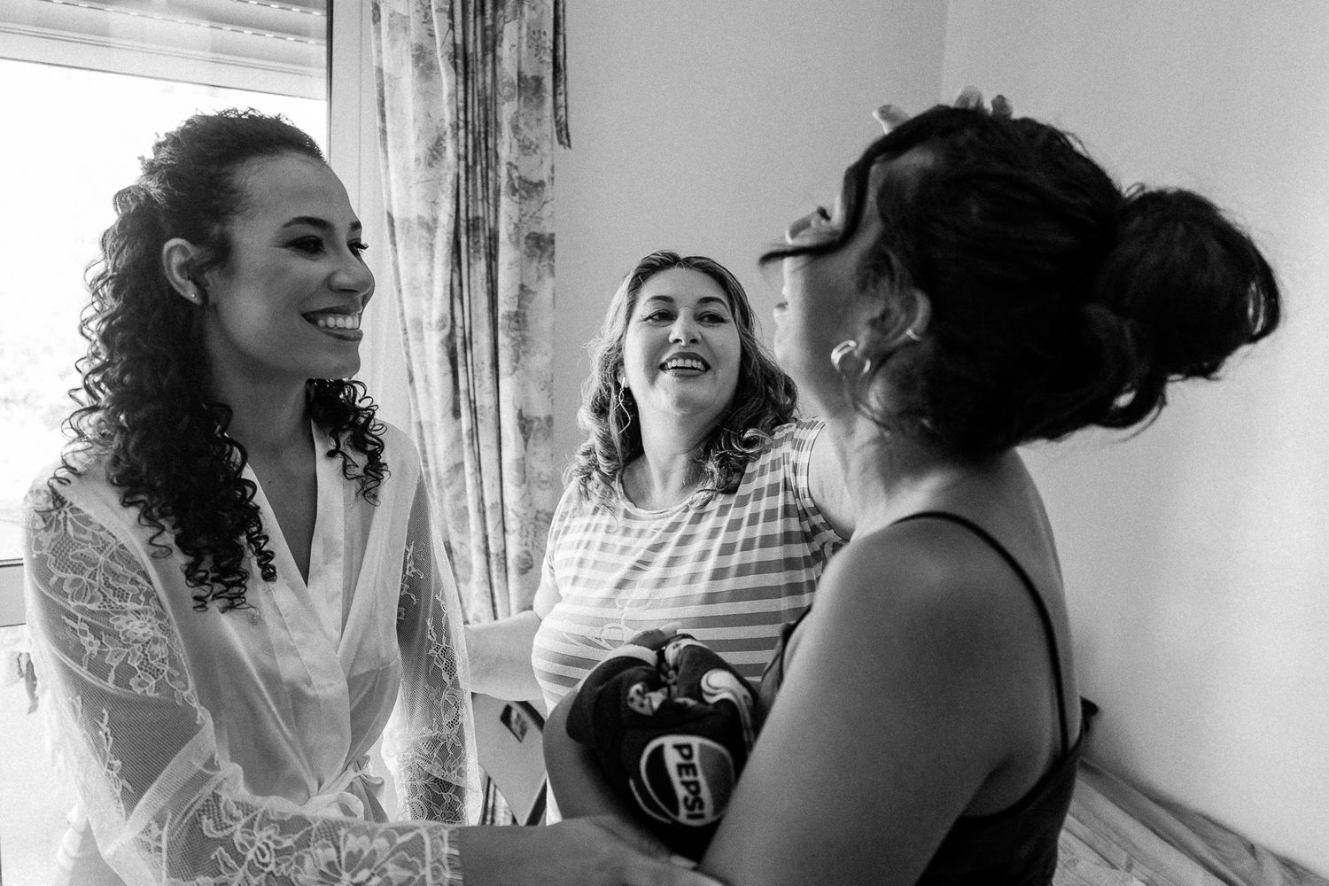 Three women laughing joyfully together in a black and white photo during wedding preparations.