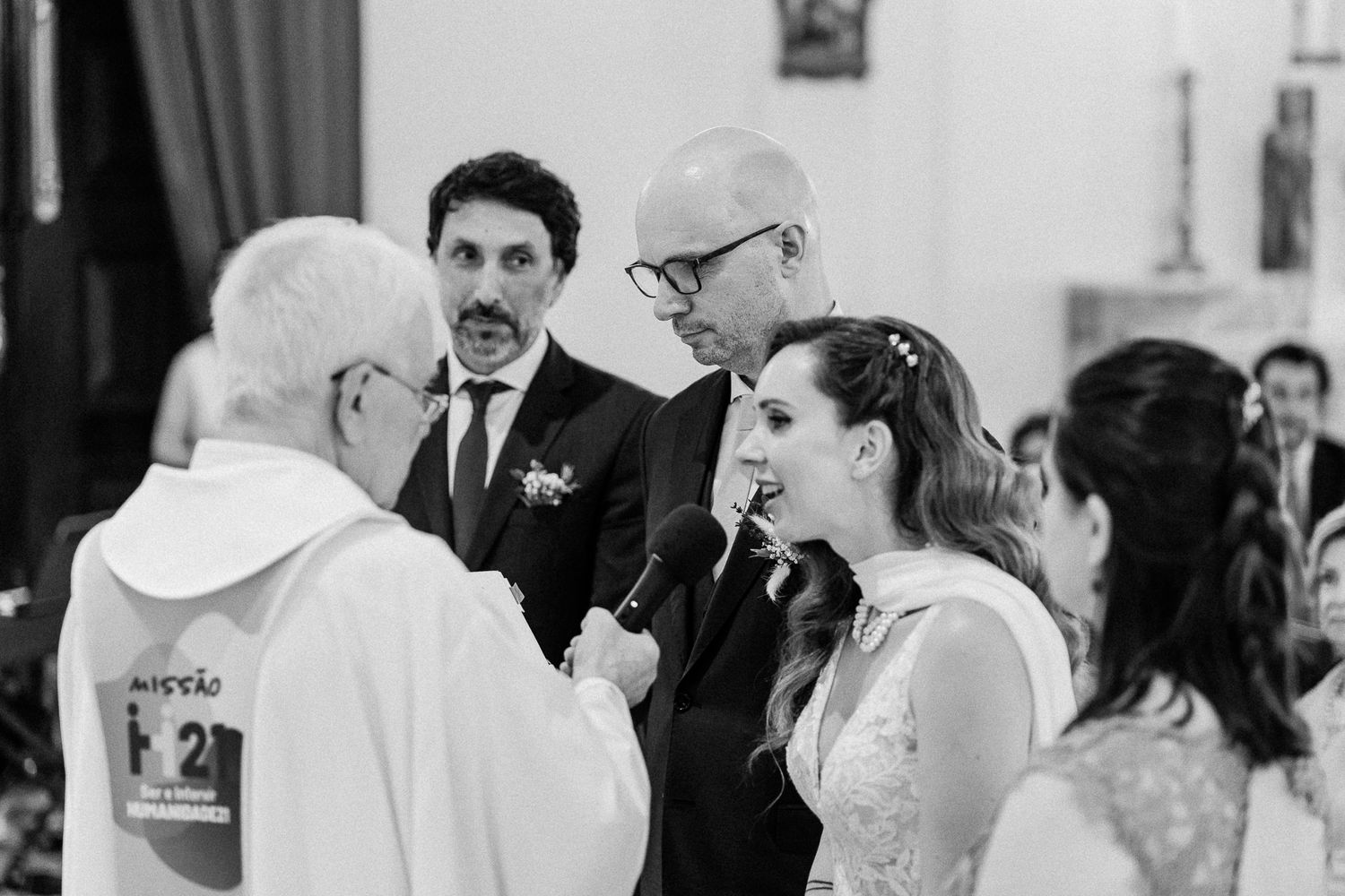 Bride and groom share a joyful moment with priest during black and white church wedding ceremony.