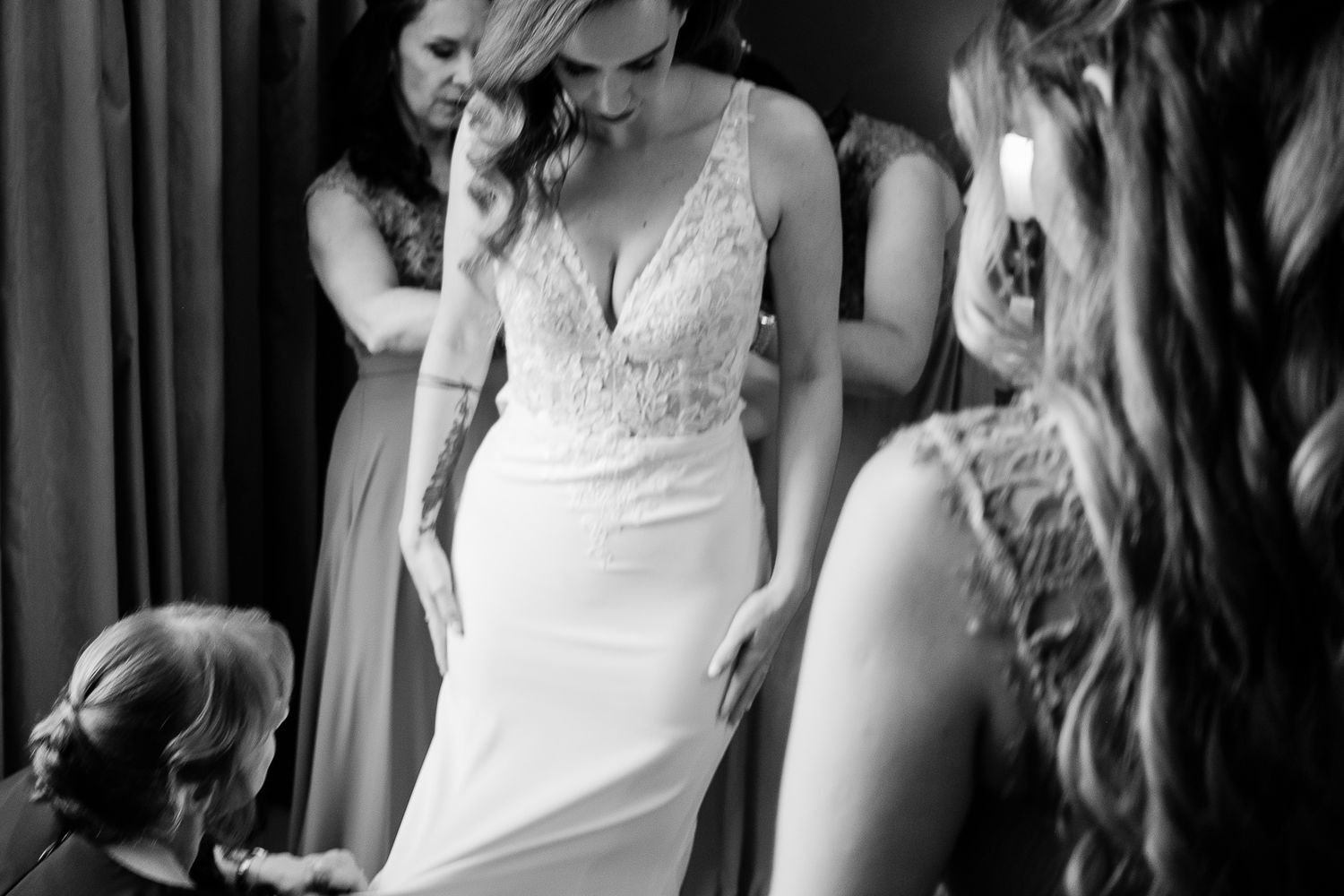 Black and white photo of bride in lace wedding gown being assisted by bridesmaids while getting ready.