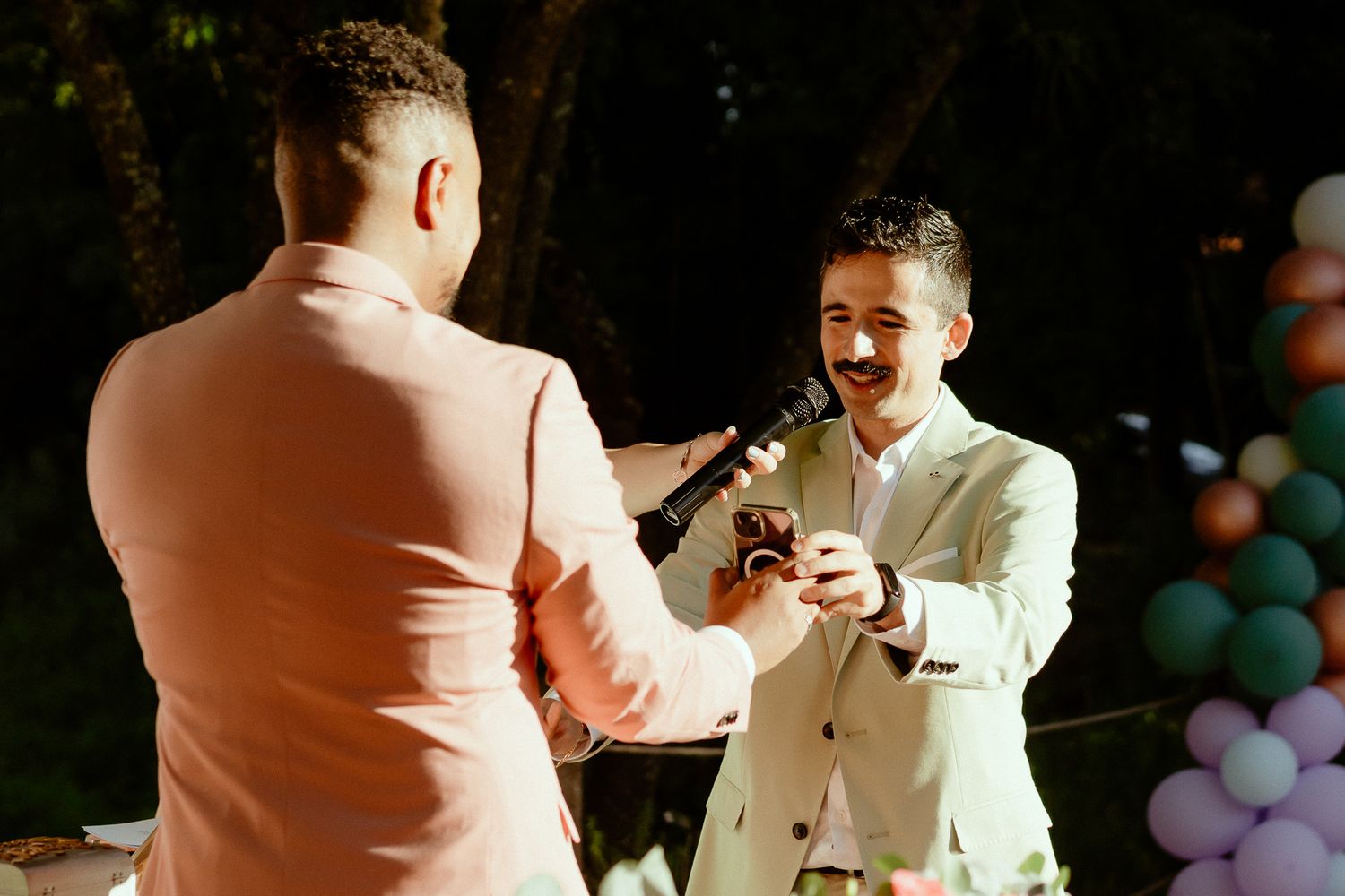 Two grooms exchange rings outdoors at a wedding ceremony, surrounded by colorful balloons.