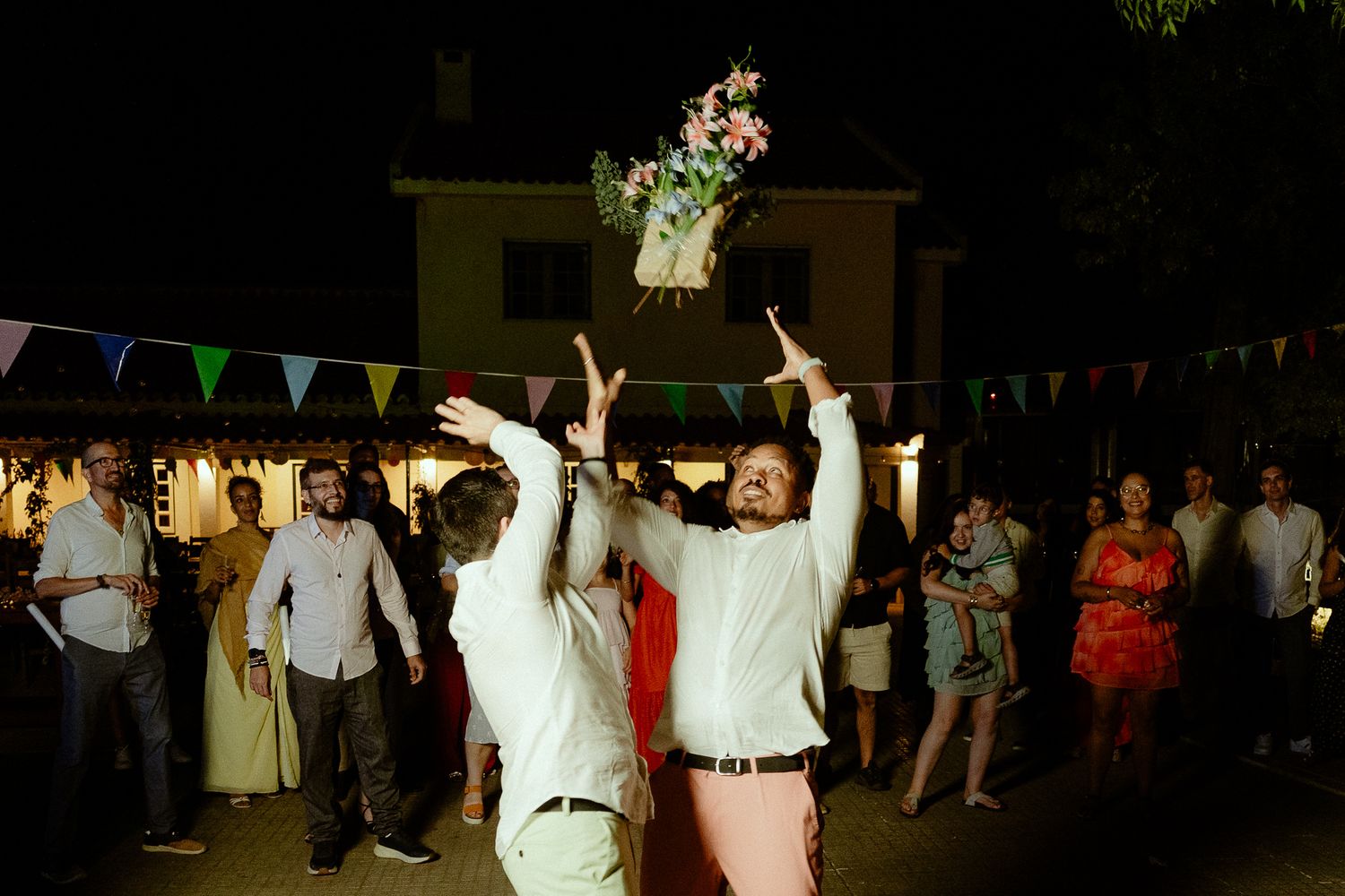 Two grooms toss a floral bouquet at an outdoor wedding reception with colorful bunting and guests.