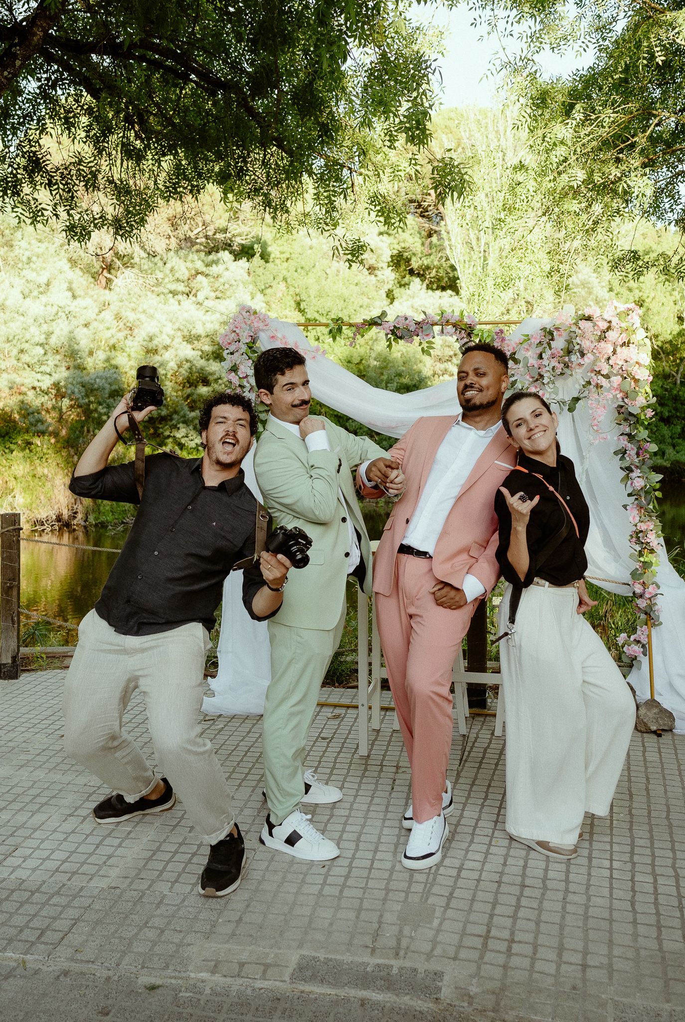 Two grooms in pastel suits pose joyfully with friends under a floral arch at an outdoor wedding ceremony.