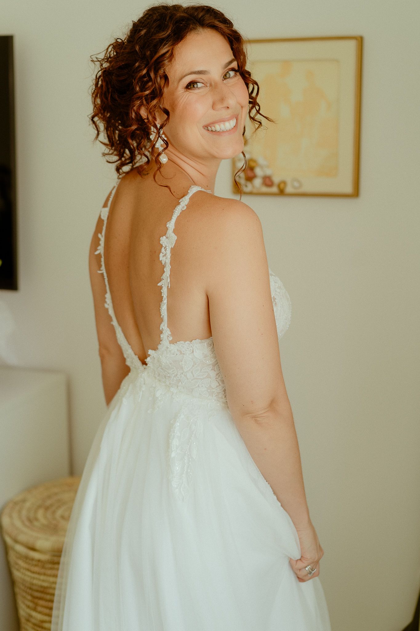 Smiling bride in a white backless lace wedding gown with spaghetti straps, showing elegant open back detail.