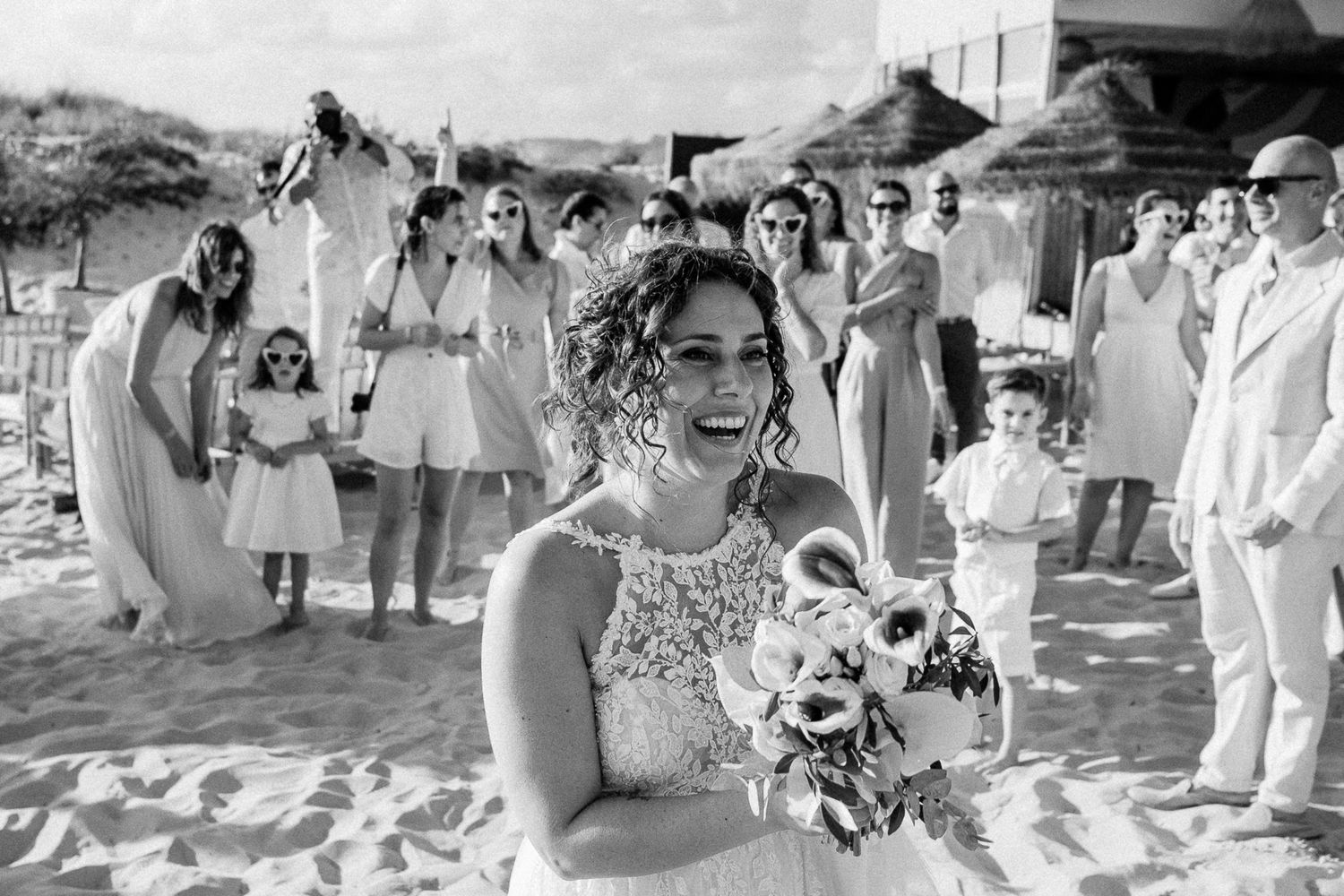 Smiling bride holding bouquet on beach wedding with guests in background, black and white photo.