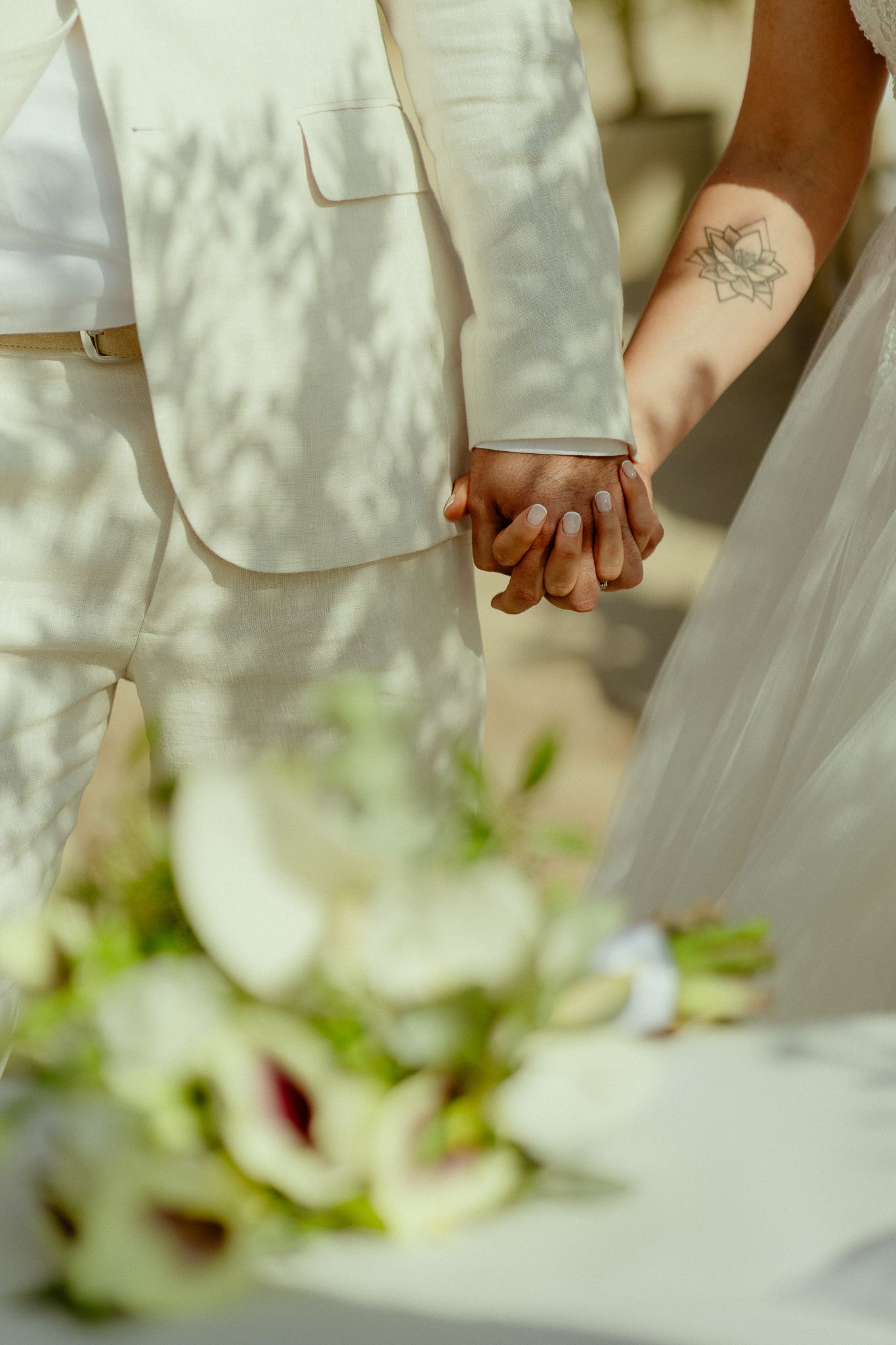 Couple holding hands at wedding, groom in white suit, bride with floral tattoo, bouquet in foreground.