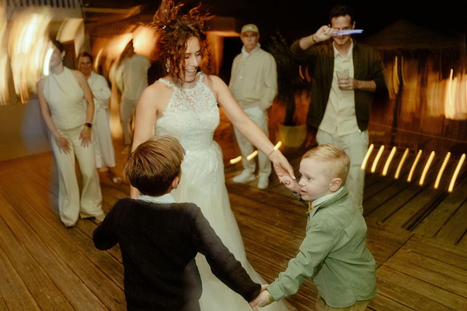 Bride in white gown dancing with two young boys on wedding reception dance floor under warm lighting.