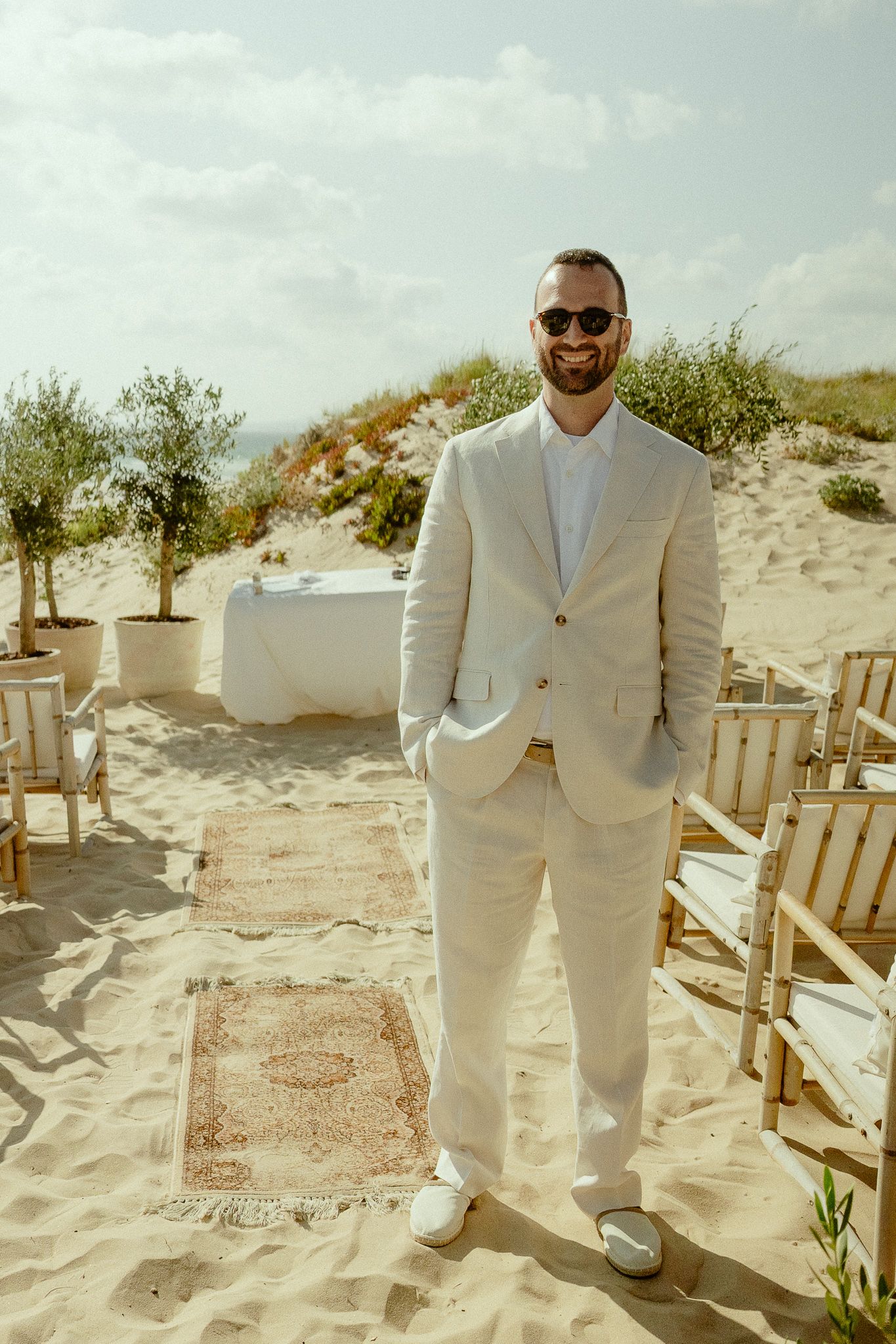 Smiling groom in white linen suit and sunglasses stands on sandy beach wedding aisle with chairs and olive trees.