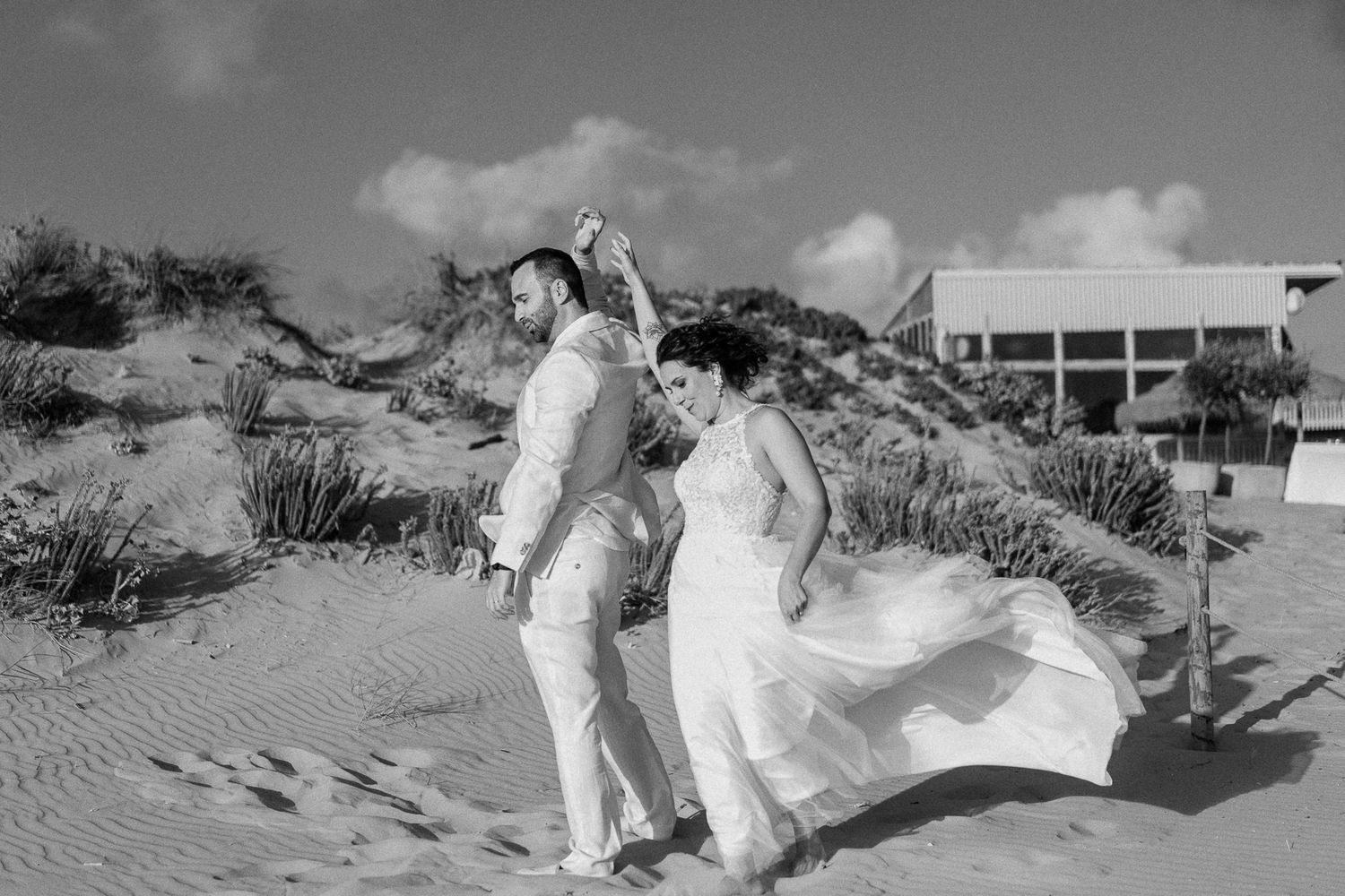 Black and white photo of a couple dancing on sandy beach dunes in wedding attire, bride's dress flowing in the wind.