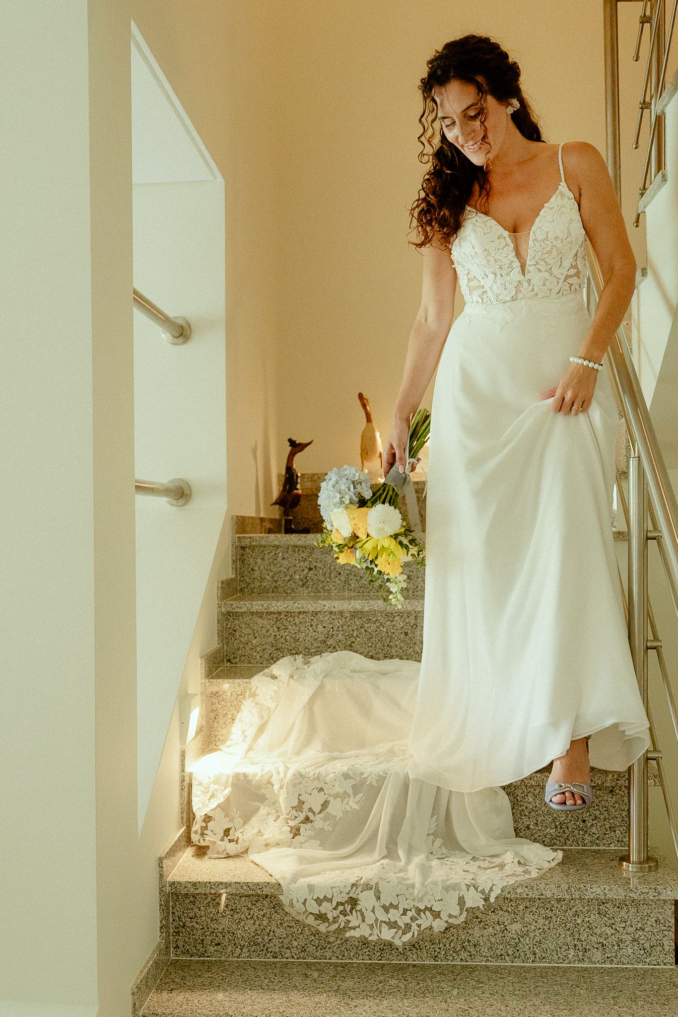 Smiling bride in white lace gown holding yellow bouquet descending marble staircase with train trailing behind her.