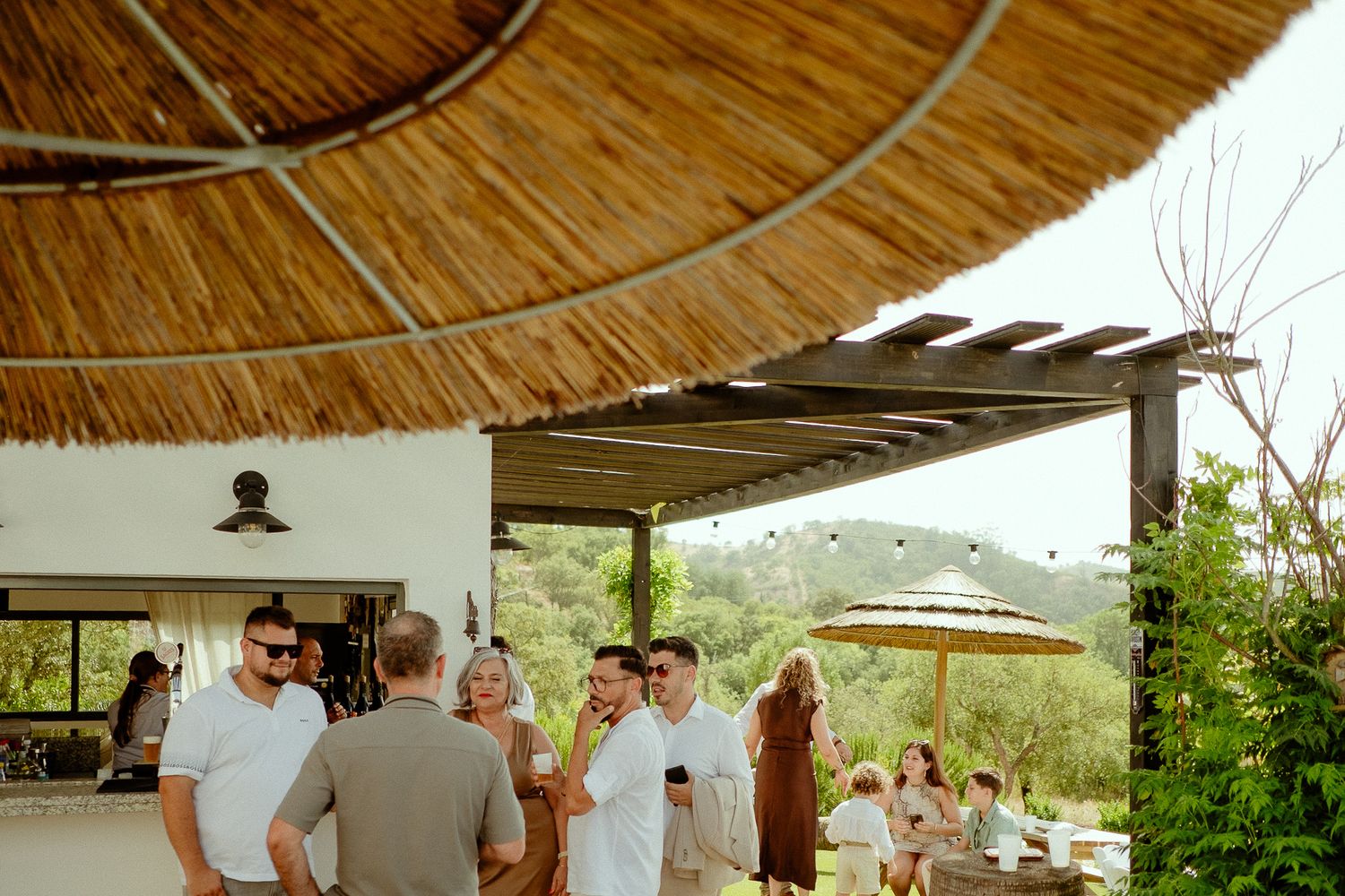 Guests mingle outdoors under a large thatched umbrella at an elegant open-air venue surrounded by lush greenery.