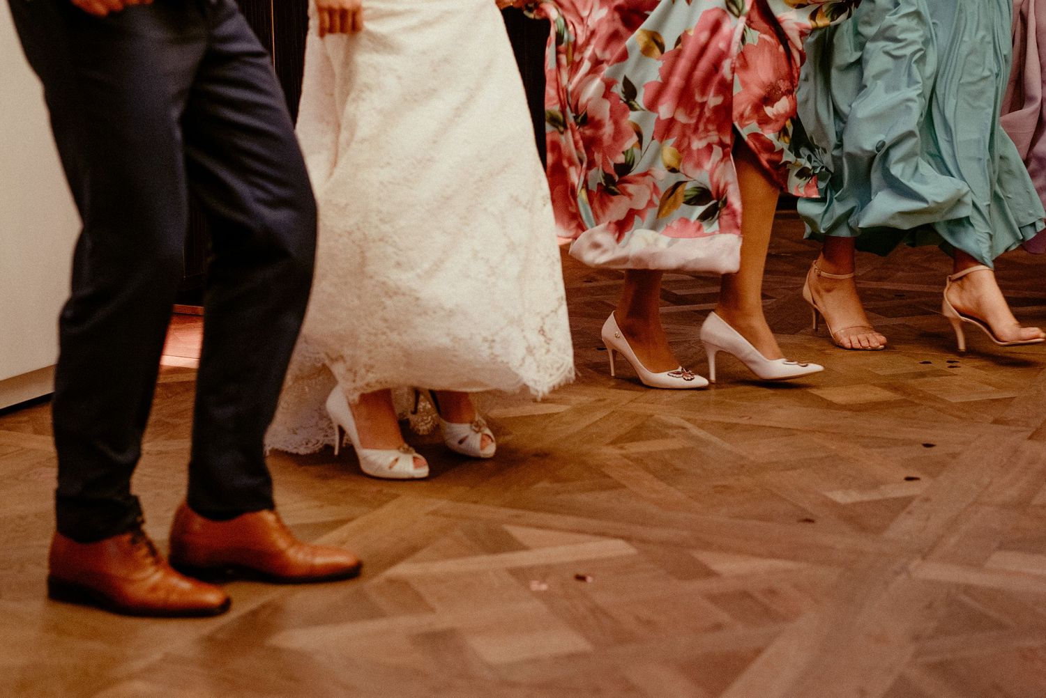 Close-up view of formal shoes and dress hemlines on a wooden dance floor at an elegant event.
