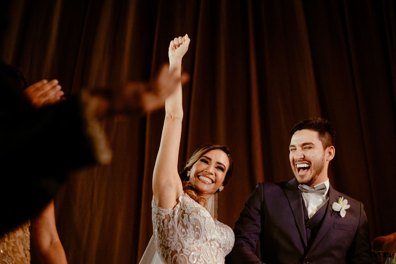 A joyful couple dances together during their wedding celebration with dark brown curtains in the background.