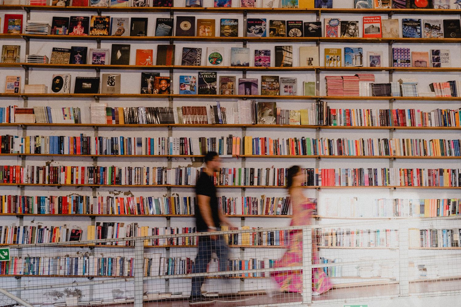Two people browsing in a bookstore with floor-to-ceiling shelves filled with colorful books.
