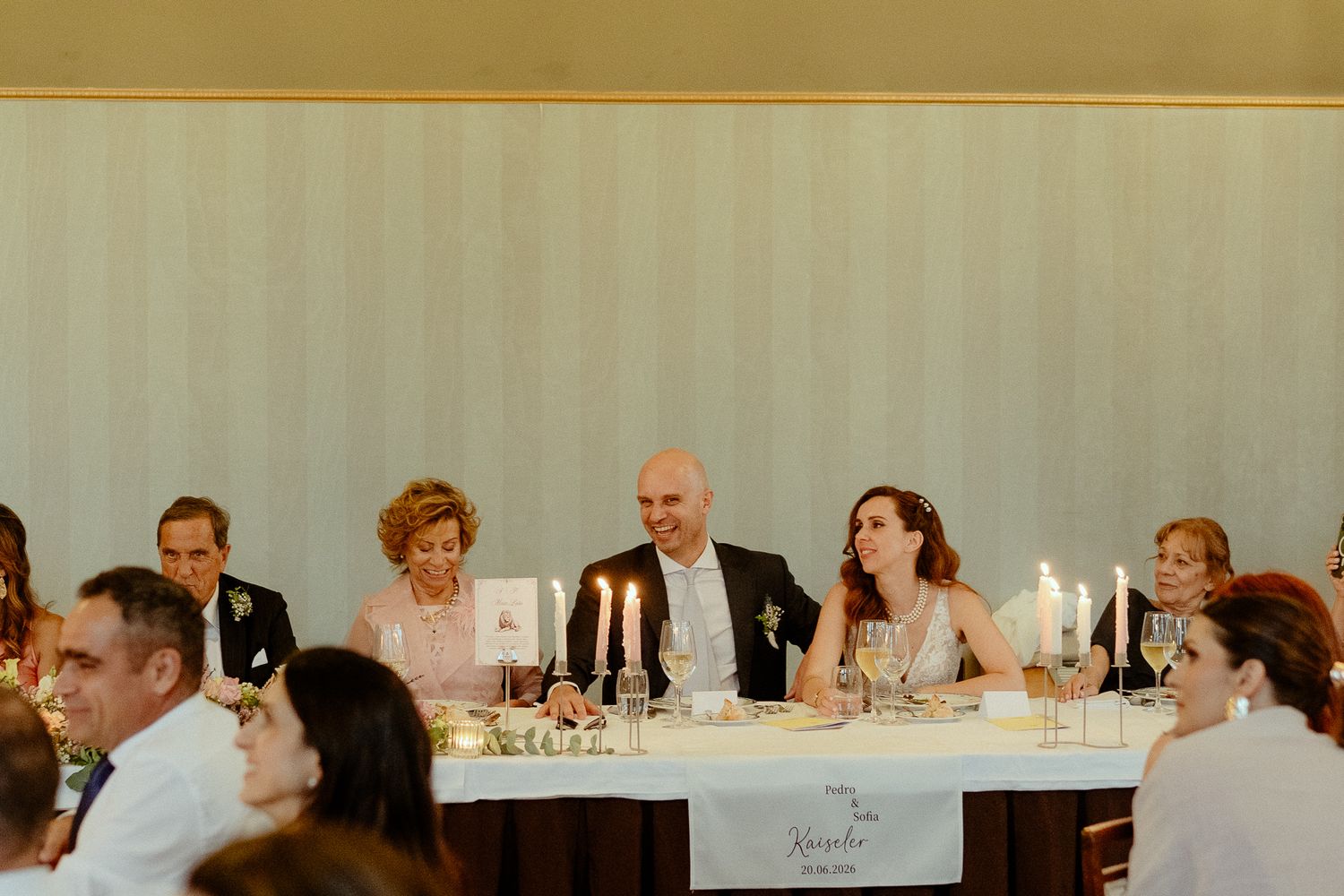 Wedding reception head table with guests seated, candles lit, elegant striped wallpaper backdrop in a formal dining room.