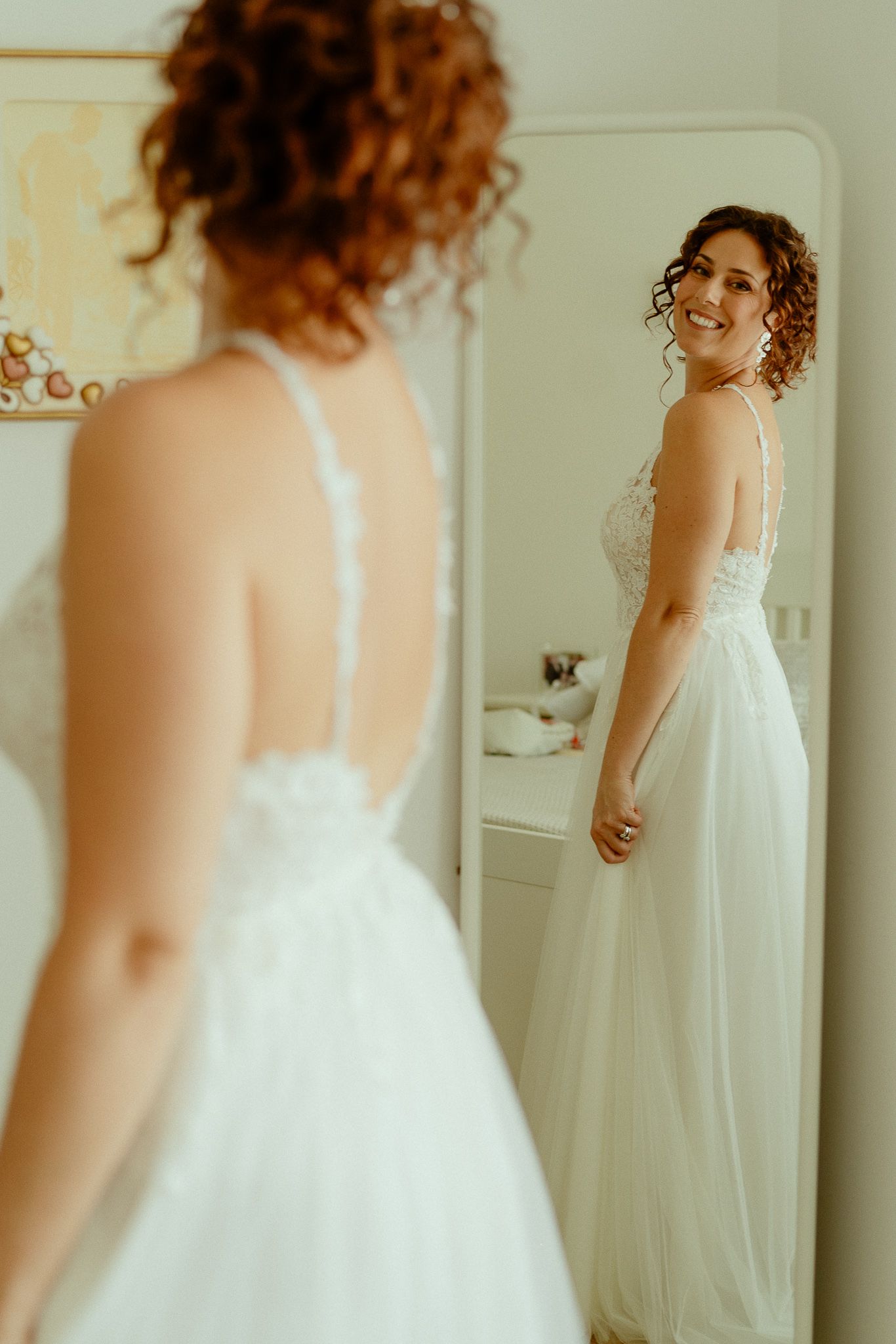 Smiling bride in a white lace backless wedding gown admiring her reflection in a full-length mirror.