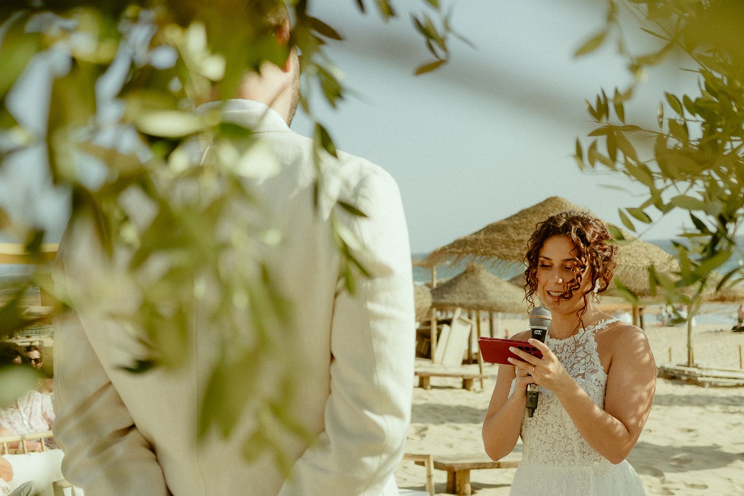 Bride in white lace dress smiles at phone during beach wedding ceremony with tropical thatched huts in background.