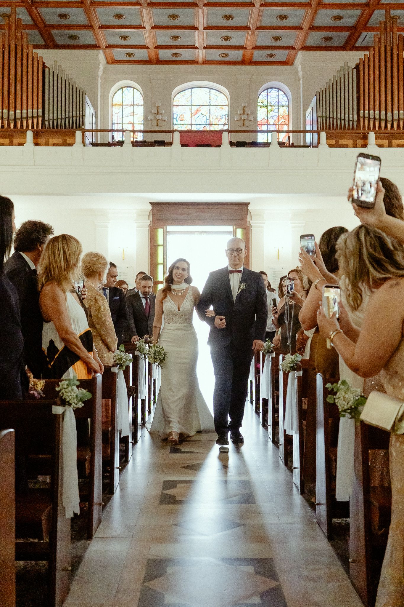 Bride walks down church aisle with escort as guests photograph the wedding processional in a classic chapel.