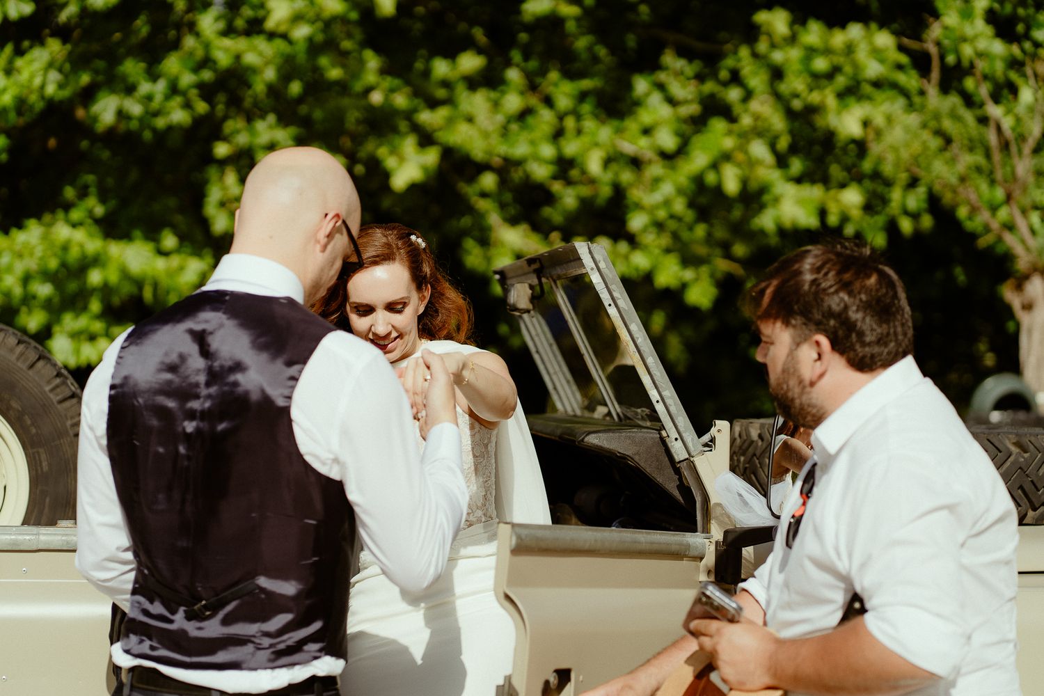 Bride and groom sharing a joyful moment beside a vintage white car with a guitarist performing outdoors.