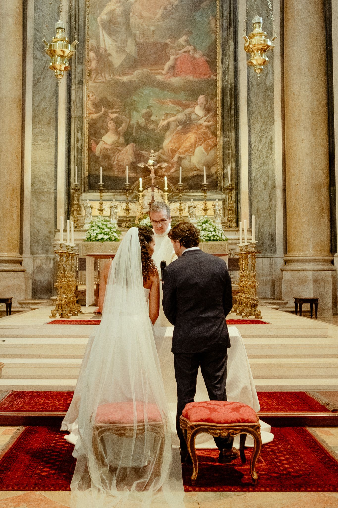 Bride and groom stand at ornate church altar during wedding ceremony, with priest officiating beneath a grand baroque painting.
